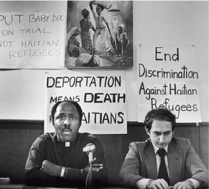 Reverend Gerard Jean Juste, (Left), and Haitian defense attorney Ira Kurzban, at deportation press conference, Miami, FL, 1981. The image is black and white and both figures are sitting in front of 3 protest posters and a painting.